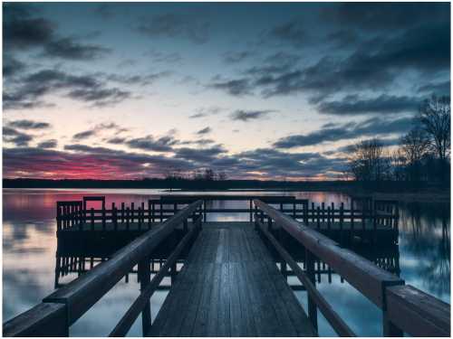 A serene lake at dusk, with a wooden dock extending into calm waters and colorful clouds reflecting in the sky.