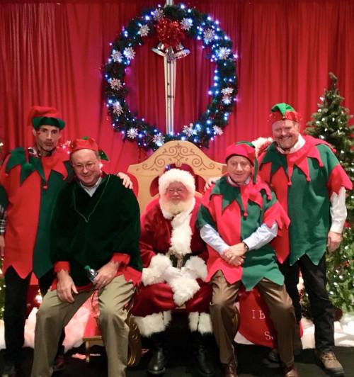 Five men dressed as elves pose with Santa Claus in front of a festive backdrop with Christmas decorations.
