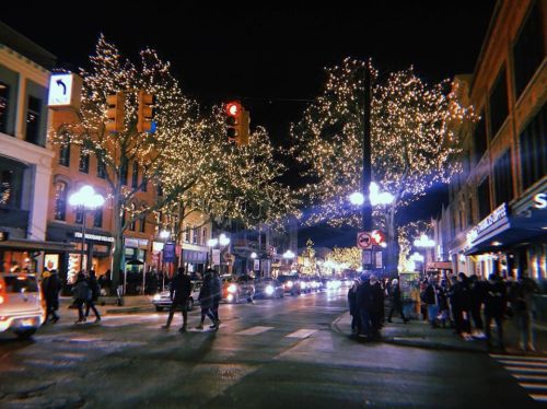 A bustling street at night, adorned with twinkling holiday lights on trees, with people and cars moving through.