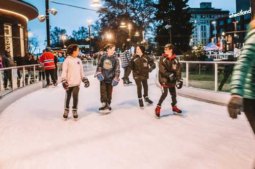 Four children skate on an ice rink, smiling and enjoying the winter atmosphere in a festive outdoor setting.