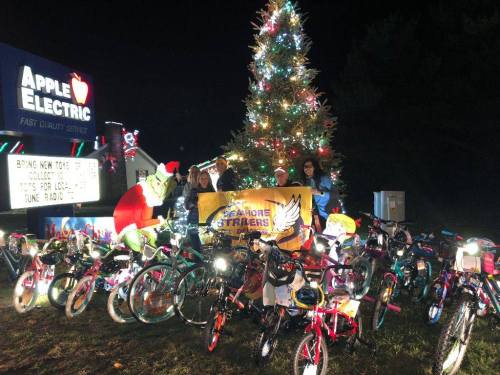 A festive scene with a decorated Christmas tree, children with bikes, and a sign promoting a toy drive event.