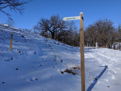 A wooden signpost points to the Sunrise Trail, surrounded by snow and bare trees under a clear blue sky.