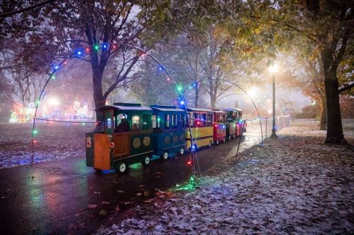A colorful train decorated with lights travels along a snowy path, surrounded by trees and festive decorations.