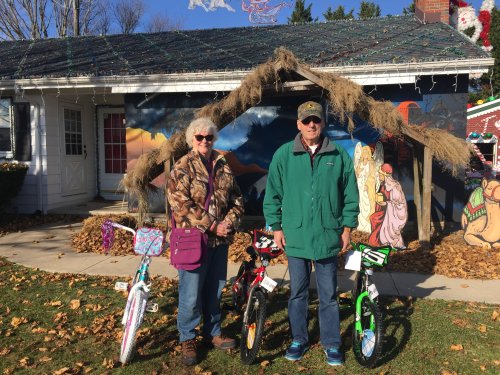 An elderly couple stands beside two colorful bicycles in front of a decorated house with a mural and festive decorations.