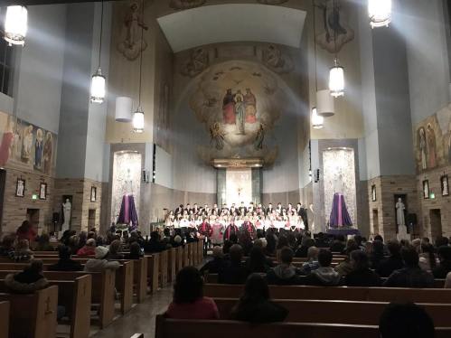 A large audience in a church, with a choir performing on stage under ornate murals and soft lighting.