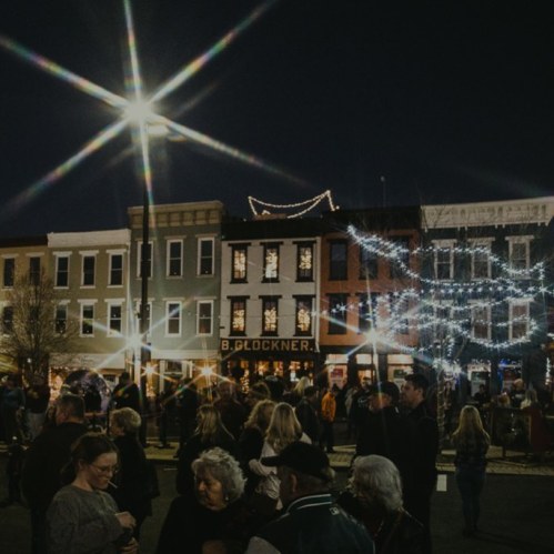 A lively street scene at night, with festive lights and crowds gathered outside shops and buildings.