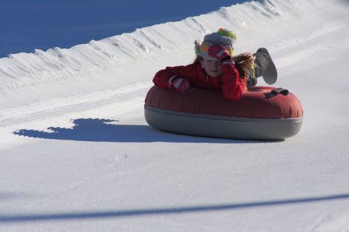 A child in a colorful outfit lies on a snow tube, enjoying a winter day on a snowy slope.