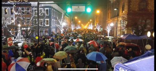 A large crowd with umbrellas gathers on a festive street, illuminated by lights and decorations.