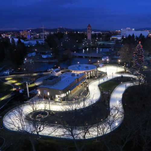 Aerial view of a winding ice skating rink surrounded by trees and a lit Christmas tree at dusk.