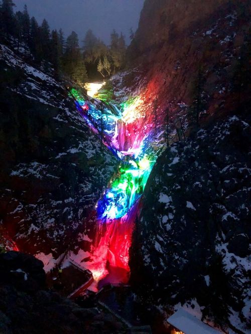 A colorful waterfall illuminated with vibrant lights, surrounded by rocky cliffs and trees in a nighttime setting.