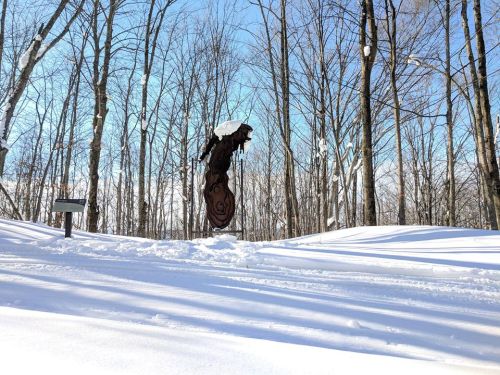 A snowy landscape with bare trees and a wooden sculpture in the foreground, under a clear blue sky.