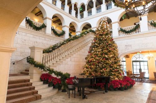 A grand staircase adorned with garlands, featuring a large Christmas tree and a piano in a festive lobby setting.