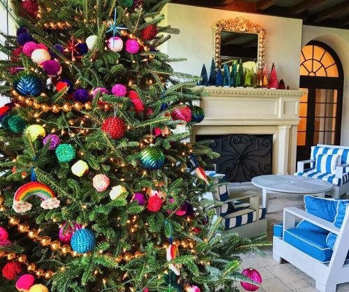 A colorful Christmas tree adorned with vibrant ornaments stands in a cozy living room with a fireplace and blue-striped chairs.