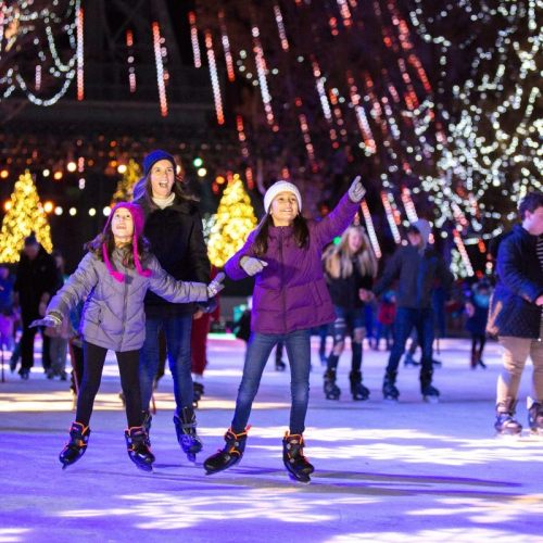 A group of people ice skating outdoors, surrounded by festive lights and holiday decorations.