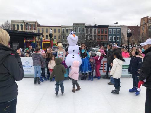 Children ice skate and gather around a snowman character, with festive buildings and a crowd in the background.