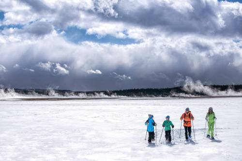 Four people in colorful winter gear snowshoeing across a snowy landscape with steam rising in the background.