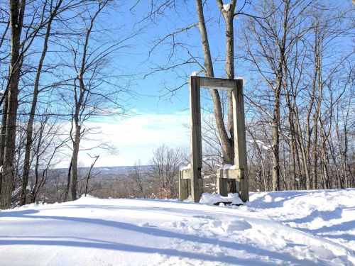 A wooden frame stands in a snowy landscape, surrounded by bare trees under a clear blue sky.