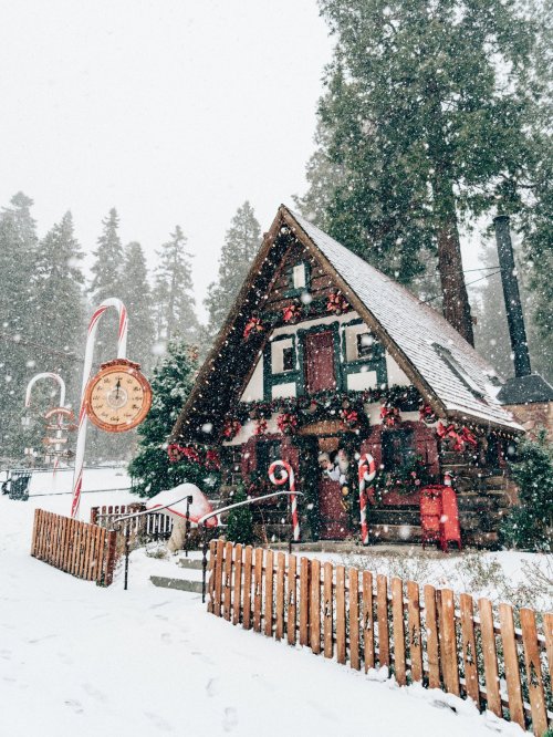A cozy, snow-covered cabin decorated for the holidays, surrounded by tall trees and falling snowflakes.