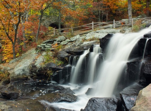 A serene waterfall cascades over dark rocks, surrounded by vibrant autumn foliage and a rustic wooden fence.