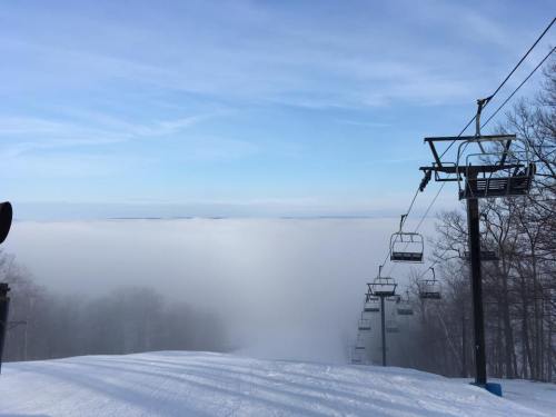A snowy ski slope with empty chairlifts and a thick fog covering the landscape below, under a clear blue sky.