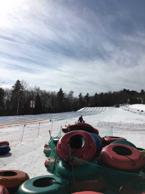 A snowy landscape with colorful snow tubes stacked in the foreground and a ski slope in the background under a cloudy sky.