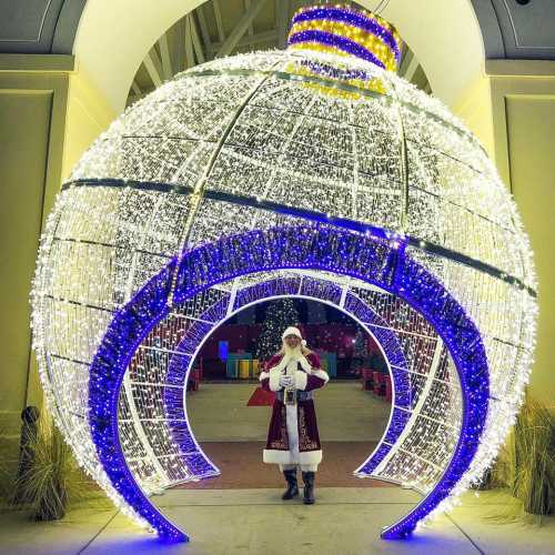 A person dressed as Santa stands inside a large, illuminated Christmas ornament display.