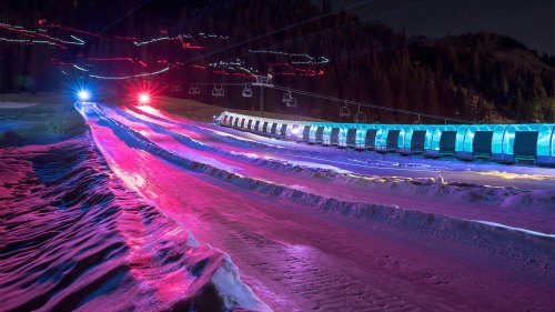 A snowy landscape at night with colorful lights illuminating a ski lift and snow-covered slopes.