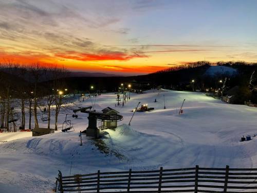 A snowy ski resort at sunset, with ski lifts and trails illuminated by lights against a colorful sky.