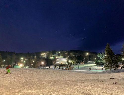 A snowy ski resort at night, illuminated by lights, with skiers on the slopes and trees in the background.