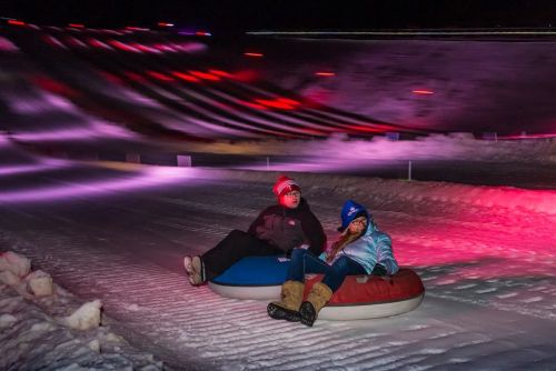 Two people sit on snow tubes on a brightly lit slope, surrounded by colorful lights in a winter setting.