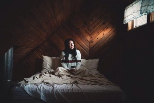 A woman sits on a bed in a cozy, dimly lit room, holding a cup and gazing thoughtfully out a window.