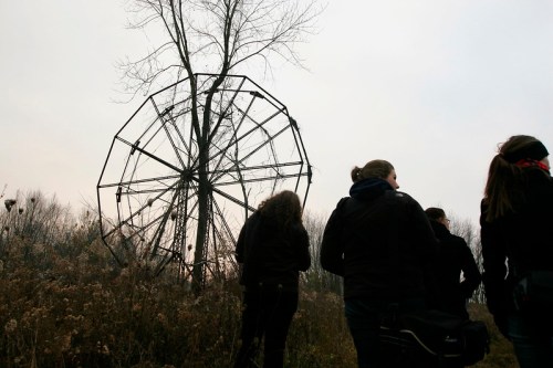 A group of people walks past a large, rusted Ferris wheel in a desolate landscape under a cloudy sky.