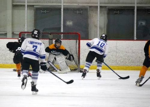 A youth hockey game with players in white and black jerseys, and a goalie in yellow, defending the net.