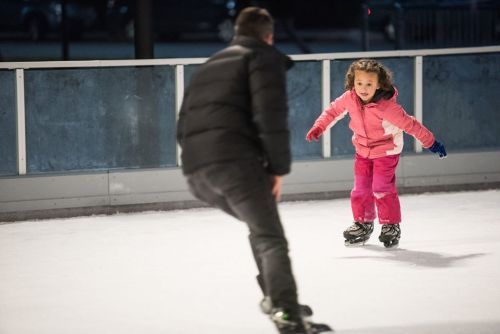 A child in a pink jacket skates joyfully on ice while an adult watches nearby.
