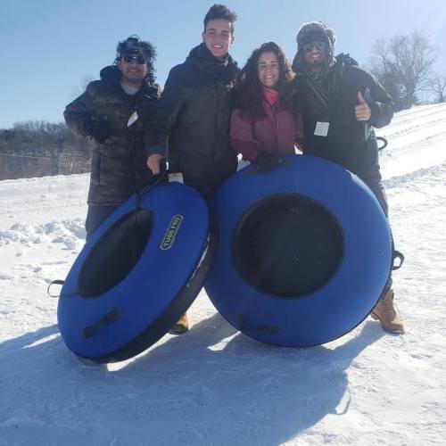 Four people stand in the snow, holding blue snow tubes, smiling in winter clothing on a sunny day.
