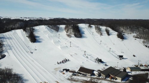 Aerial view of snow-covered hills with ski slopes and a small building at the base, surrounded by trees.