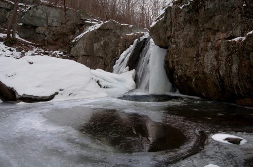 A frozen waterfall surrounded by icy rocks and snow, with a calm pool of water reflecting the scene.