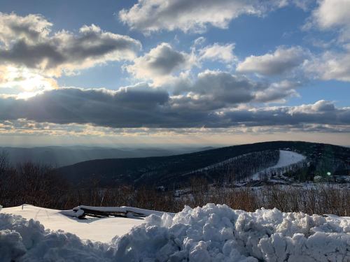 A snowy landscape with rolling hills under a cloudy sky, illuminated by the sun peeking through the clouds.