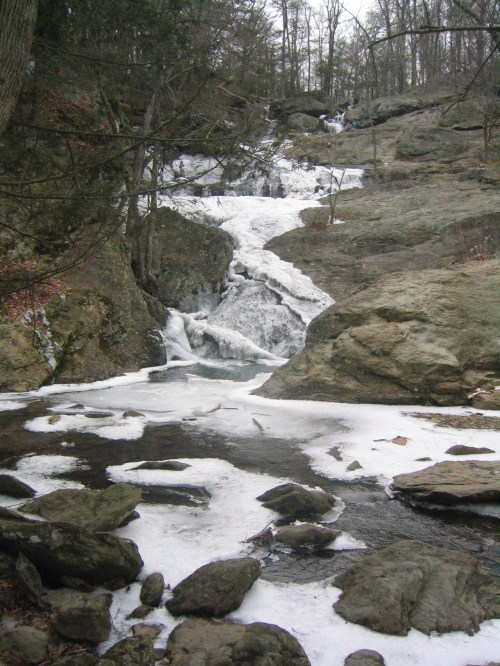 A frozen waterfall cascades over rocky terrain, surrounded by bare trees and patches of ice.