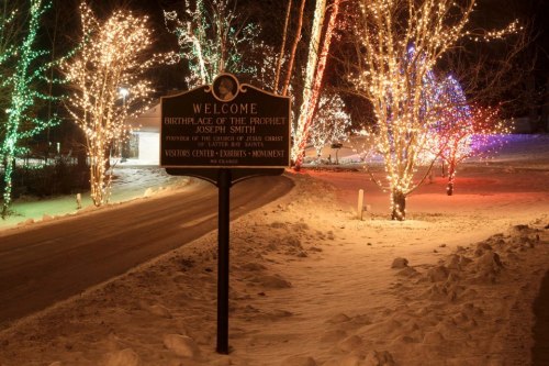 A snowy scene featuring a sign that reads "Welcome: Birthplace of the Prophet Joseph Smith," surrounded by colorful lights.