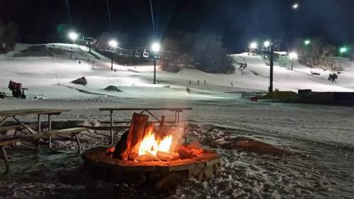 A cozy fire pit surrounded by snow, with ski slopes and lights in the background at night.