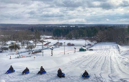 A snowy hill with six people sitting on sleds, ready to go tubing, surrounded by trees and a winter landscape.