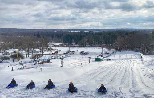 A snowy hill with six people sitting on sleds, ready to go tubing, surrounded by trees and a winter landscape.