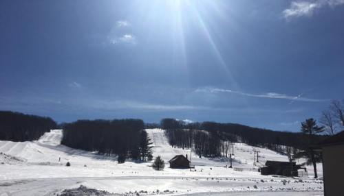 Snow-covered ski slopes under a bright blue sky with a shining sun and distant trees.