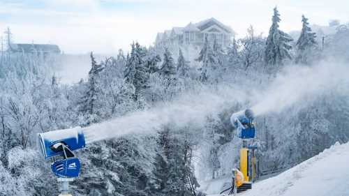 Snowmaking machines spray artificial snow in a winter landscape with frosted trees and a distant building.