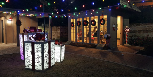 Festive outdoor scene with illuminated gift boxes, holiday lights, and decorated windows.