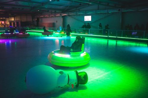 A colorful indoor rink with glowing bumper cars and a fallen snowman, surrounded by people enjoying the scene.