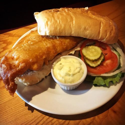 A plate featuring a crispy fish sandwich with lettuce, tomato, pickles, and a side of tartar sauce.