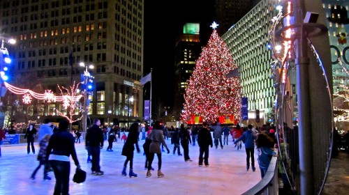 A festive ice skating rink at night, featuring a large, decorated Christmas tree and colorful lights in the background.