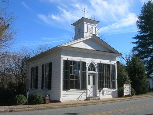 A white church building with a cross on top, large windows, and a sign outside, set against a clear blue sky.